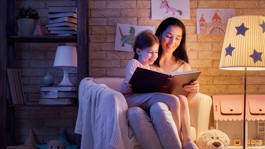 Le fasce orarie per risparmiare energia. Mamma che legge fiabe alla bambina sotto la luce fiocca di una lampada.